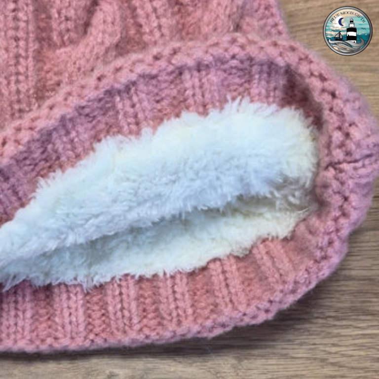 A close-up of a pink cable-knit beanie hat showing a soft white faux-fur lining inside the ribbed cuff, placed on a light wood surface.