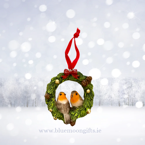 A Christmas ornament shaped like a green pine wreath with two robin birds in the center and a red hanging ribbon, shown against a snowy winter scene with blurred lights. The text 'www.