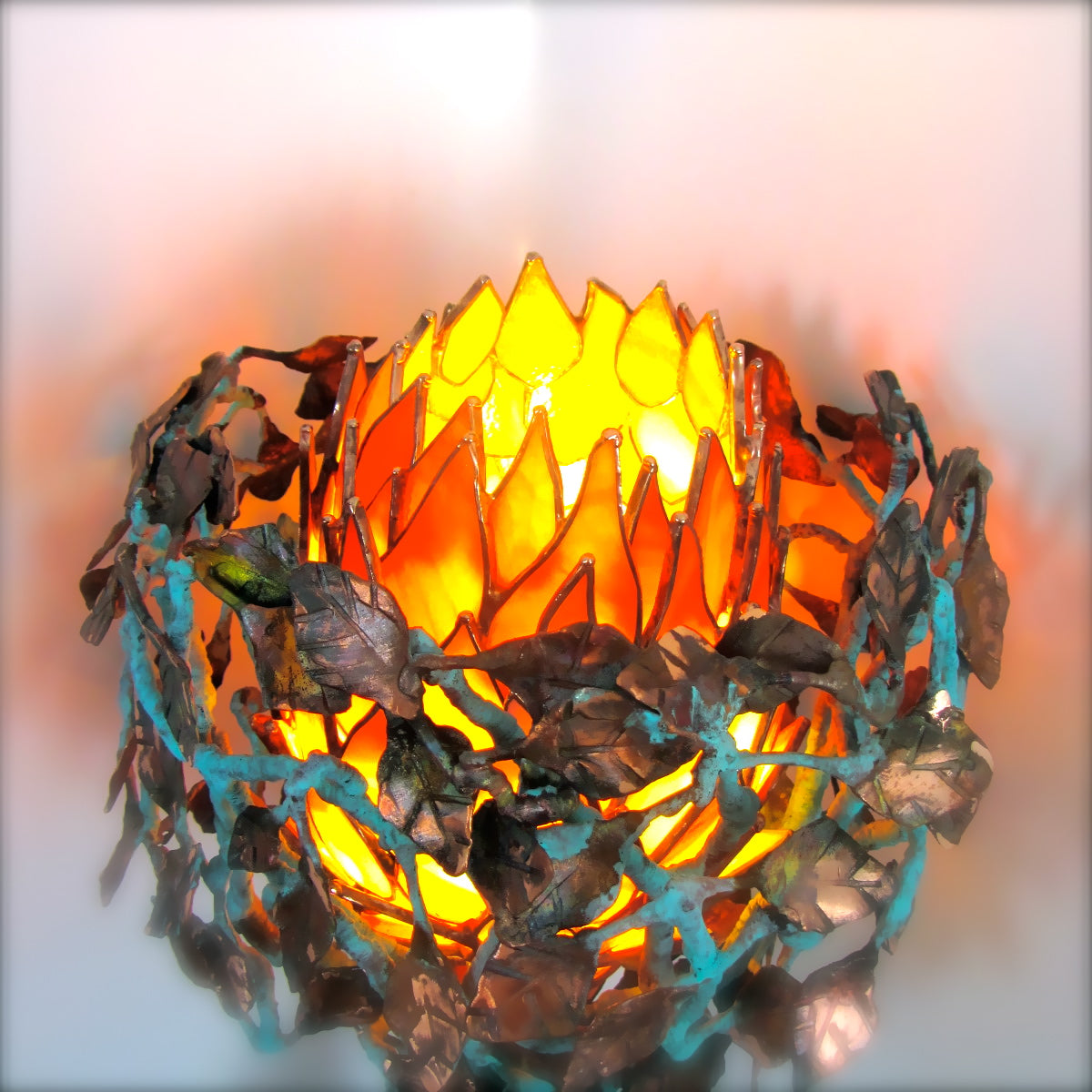 A decorative candle holder featuring a glowing orange and yellow stained-glass flower center surrounded by a ring of dark metallic leaf cutouts and turquoise blue vine accents, shown against a soft blurred light background.