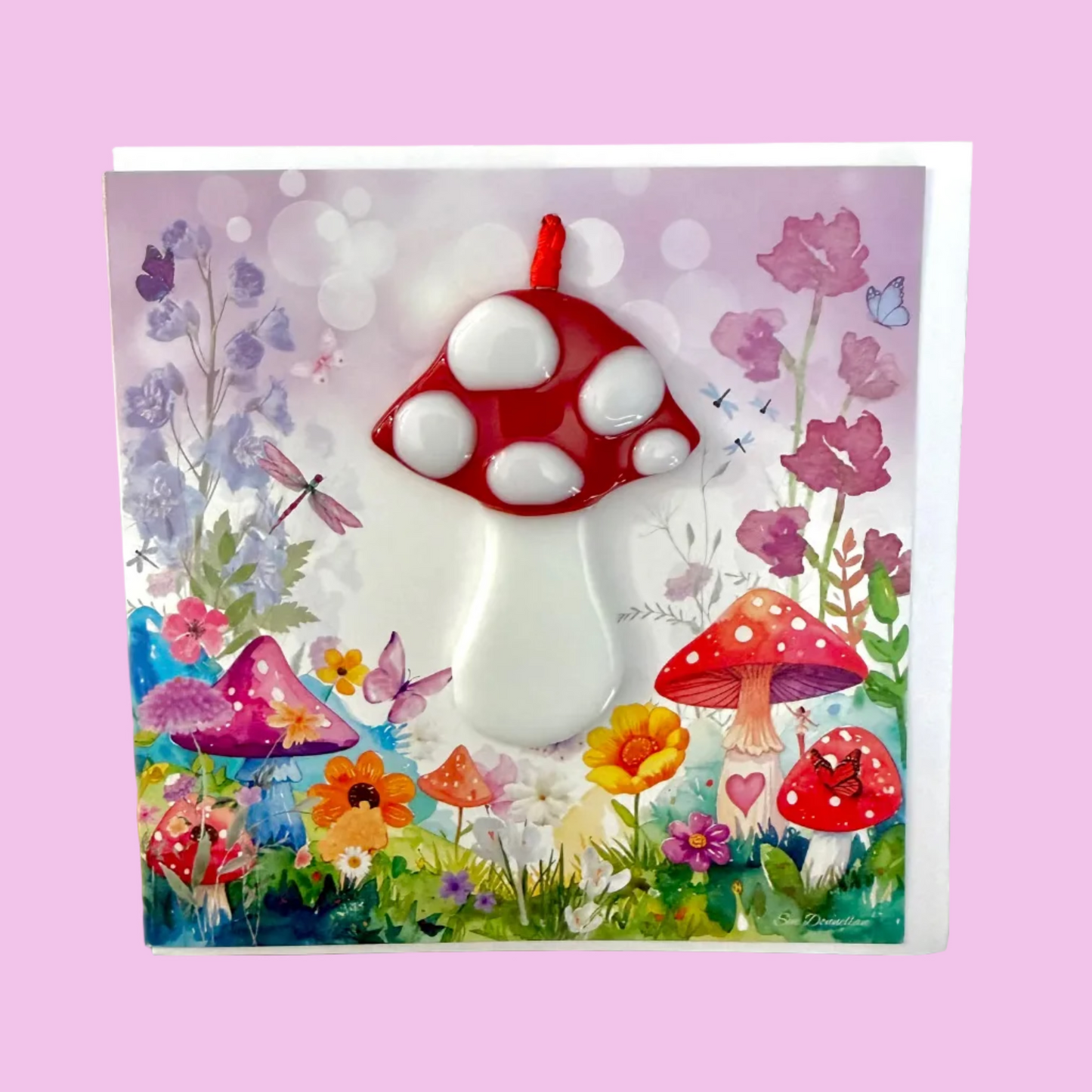 A square decorative card or art print featuring a whimsical mushroom garden scene with a large red-and-white mushroom in the center, smaller mushrooms, flowers, butterflies, and a soft pastel background, photographed on a light wood surface.
