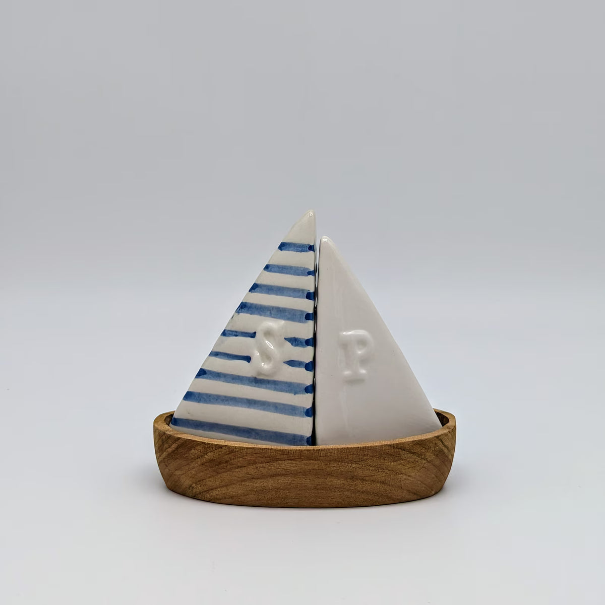 A small decorative sailboat-shaped object with two triangular sails mounted on a wooden base. One sail is white with blue stripes and a raised letter 'S', and the other is white with a raised letter 'P'.