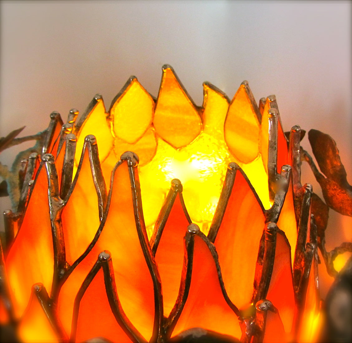 A close-up of a decorative stained-glass candle holder or lamp with a glowing warm yellow and orange flame-like design, outlined in dark metal with small leaf-like accents.