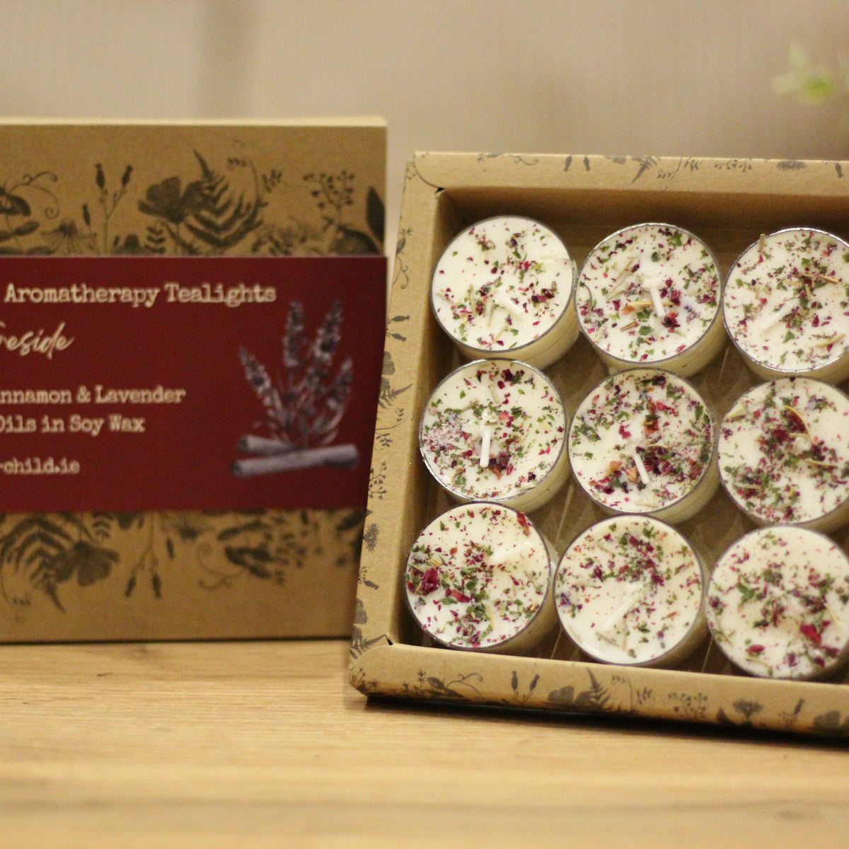 A box of handmade aromatherapy tea lights shown next to its open packaging on a wooden surface. The tea lights are round white candles topped with dried flower petals, and the kraft paper box has a dark red label with botanical illustrations.