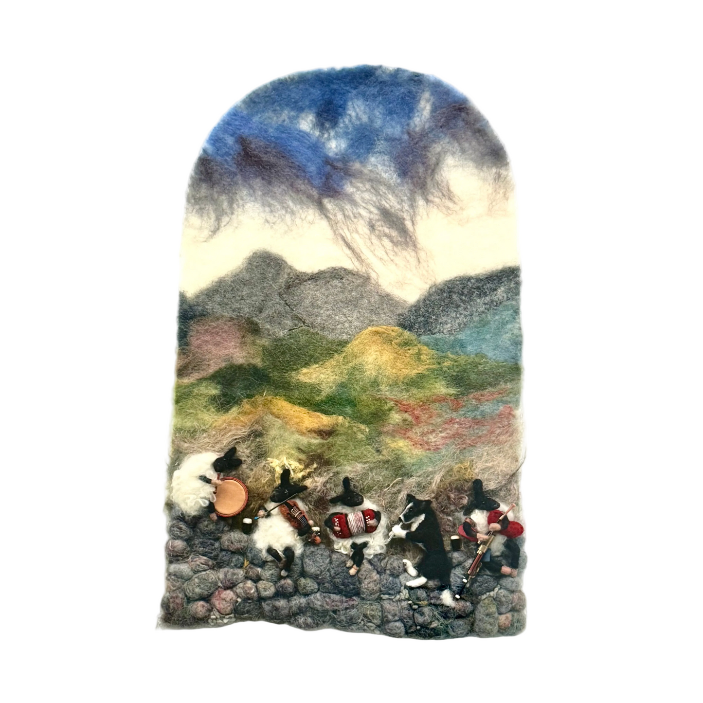 A framed art piece featuring a felted wool landscape with small black-and-white dogs playing musical instruments, displayed on a wooden surface with jewelry boxes in the background.