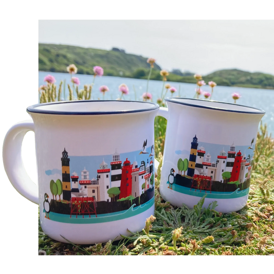 Two white enamel mugs with a colorful lighthouse and harbor illustration, placed on grass outdoors near water and flowers.