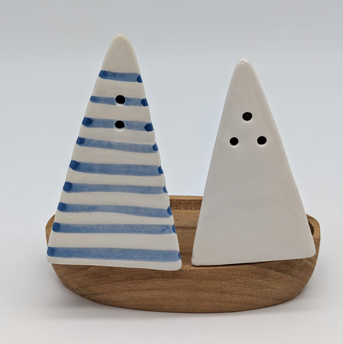 A decorative salt and pepper shaker set shaped like a small sailboat, featuring a blue-and-white striped conical figure and a plain white conical figure mounted on a rounded wooden base.