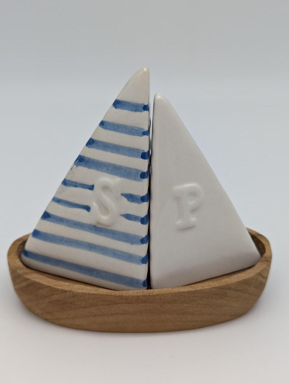 A decorative ceramic sailboat-shaped salt and pepper shaker set on a wooden base. One sail is white with blue horizontal stripes and a raised letter 'S', while the other is solid white with a raised letter 'P'.